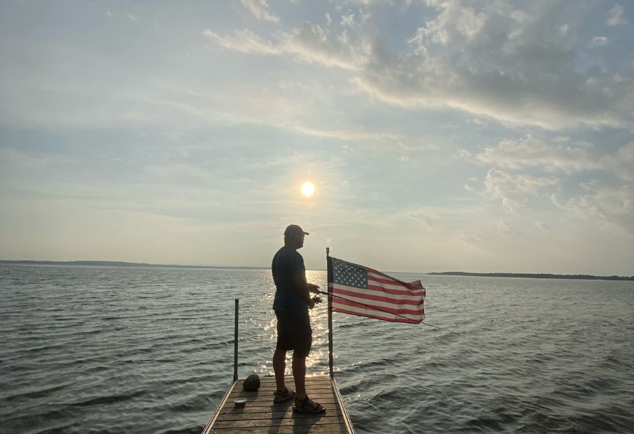 Man fishes off a dock overlooking Lake Champlain with an American flag on his right.